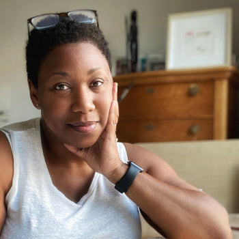 Woman sitting on a couch with her hand on her chin, wearing a white tank top and black watch.